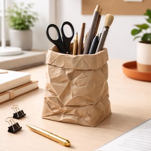 Op de afbeelding: Een bruine bureau-organizer in papieren zakstijl gevuld met zwarte schaar, pennen en een make-upkwast. Een gouden pen, zwarte paperclips en een open boek liggen op het lichte houten bureau. Een witte bureaulamp en een plant in een pot staan op de achtergrond.