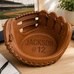 May include: A brown baseball glove shaped bowl featuring a textured leather-like surface with visible lacing details. The interior of the bowl is embossed with the name JACKSON and the number #12 in a bold, capitalized sans-serif font style.