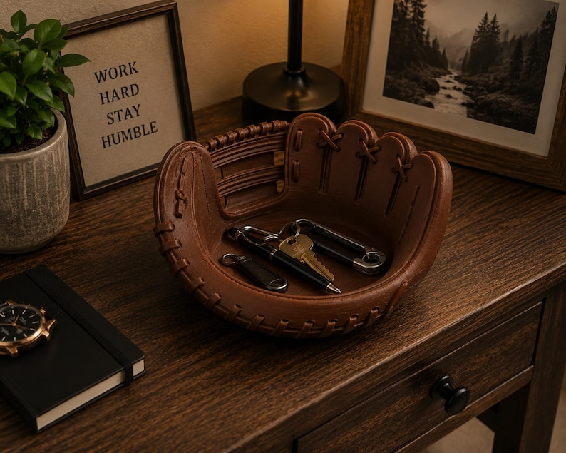 K&ouml;nnte beinhalten: Eine braune, lederne Baseballhandschuh-f&ouml;rmige Ablageschale enth&auml;lt Schl&uuml;ssel und einen Stift. Die Schale steht auf einer Holzoberfl&auml;che neben einem gerahmten Bild und einer kleinen Topfpflanze. Der gerahmte Text lautet "WORK HARD STAY HUMBLE."