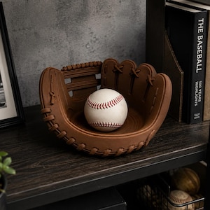 K&ouml;nnte beinhalten: Eine braune, baseballhandschuhf&ouml;rmige Schale enth&auml;lt einen wei&szlig;en Baseball mit roten N&auml;hten. Die Schale steht auf einem dunklen Holzregal, neben einem gerahmten Schwarz-Wei&szlig;-Foto eines Baseballspielers und Baseballb&uuml;chern.