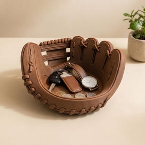 May include: A brown baseball glove-shaped catch-all dish holds keys, a watch, and coins. The dish is made of a textured material and has detailed stitching. A small plant in a pot is in the background.