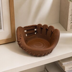 K&ouml;nnte beinhalten: Eine braune, baseballhandschuhf&ouml;rmige Dekoschale. Die Schale hat detaillierte N&auml;hte und Schn&uuml;rungen. Die Schale besteht aus einem festen Material und steht auf einem wei&szlig;en Regal.