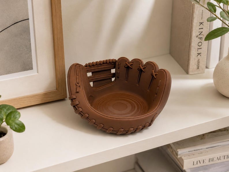 K&ouml;nnte beinhalten: Eine braune, baseballhandschuhf&ouml;rmige Dekoschale. Die Schale hat detaillierte N&auml;hte und Schn&uuml;rungen. Die Schale besteht aus einem festen Material und steht auf einem wei&szlig;en Regal.