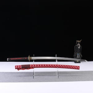 Könnte beinhalten: Ein japanisches Katana-Schwert mit einem rot-schwarzen Griff und einer silbernen Klinge, ausgestellt auf einem transparenten Ständer. Die Schwertscheide ist rot mit einem geometrischen Muster. Eine Miniatur-Samurai-Figur befindet sich im Hintergrund.