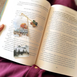 Può includere: Un segnalibro stravagante con un'auto d'epoca con bagagli, una scena di foresta e un ciondolo dorato. Il ciondolo ha un emblema colorato. Il segnalibro è in un libro aperto.