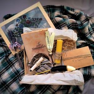 May include: A themed gift box featuring a framed floral print, a journal with the words "So remember", a candle, a feather quill, and a handwritten note. The box is lined with white paper and rests on a green and blue plaid blanket.