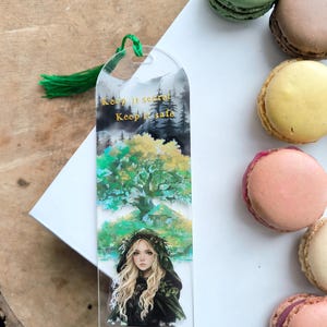 Può includere: Segnalibro in acrilico trasparente con nappa verde. Il segnalibro presenta una scena di foresta con una donna con un mantello con cappuccio, il testo "Keep it secret, Keep it safe" in lettere dorate. Macarons colorati sullo sfondo.