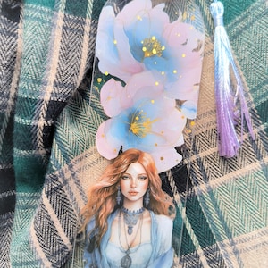 May include: A clear acrylic bookmark with a watercolour illustration of a woman with long red hair and a blue dress. The bookmark also has a floral design in shades of blue, pink, and gold, with a blue tassel.