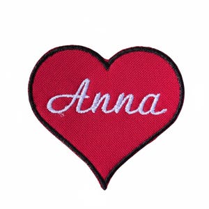 May include: A red heart-shaped embroidered patch with a black border. The name "Anna" is written in white cursive script across the centre. The patch is likely made of fabric and designed for application on clothing or accessories.