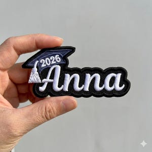 May include: Embroidered patch featuring the name "Anna" in white script on a black background. A small graduation cap with the year "2026" is above the name. The patch is held in a hand against a light grey background.