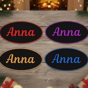 May include: Four embroidered oval patches, each with the name "Anna" in a different colour: red, purple, gold, and blue. They have a black background and a coloured border. The patches are displayed on a wooden surface.