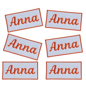 May include: Six white rectangular patches embroidered with the name "Anna" in orange cursive script. Each patch has an orange border. The patches are arranged in two rows of three.