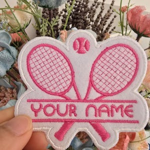 May include: White patch featuring pink embroidered tennis rackets and a tennis ball. The text "YOUR NAME" is embroidered in pink. The patch is held in front of a floral arrangement.