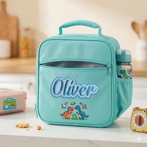 May include: A teal lunch bag with the name "Oliver" embroidered on the front. The bag has a zippered pocket and a side pocket holding a water bottle. Two cartoon dinosaurs and rockets decorate the front. A small timer and a pink lunchbox are also visible.