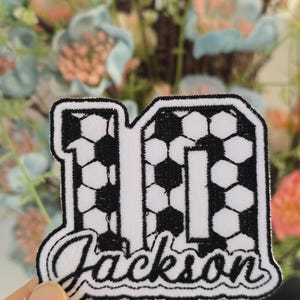 May include: An embroidered patch with the number 11, filled with a black and white soccer ball design. The name "Jackson" is written in cursive below. The patch has a black border.