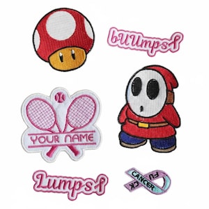 May include: A selection of embroidered patches with diverse designs. Includes a red and white mushroom, a Shy Guy character, a tennis racket design with the text "YOUR NAME", and patches with the words "bumps", "lumps", and a ribbon with "Cancer Fu".