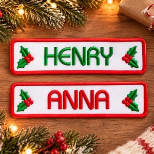 May include: Two rectangular embroidered patches with white backgrounds and red borders. The top patch features the name HENRY in green letters, and the bottom patch features the name ANNA in red letters. Both patches include green holly leaves and red berries.