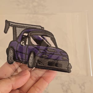 May include: An embroidered patch featuring a purple sports car with a large rear wing, a dark grey front bumper, and silver wheels. The car is depicted in a stylised manner with white windscreen accents, all contained within a clear plastic sleeve.