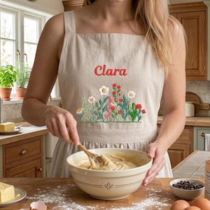 Op de afbeelding: Beige schort met geborduurd bloemenontwerp en de naam "Clara" in het rood. Het schort wordt gebruikt in een keuken, met een mengkom en ingrediënten zichtbaar. Het schort is gemaakt van een natuurlijke stof.