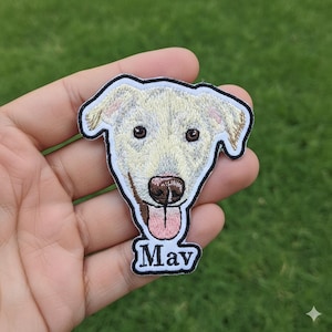 May include: Embroidered patch depicting a cream-coloured dog's face with brown eyes and a pink tongue. The patch has a black border and the name "Mav" in white script below. Suitable for clothing or accessories.