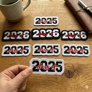 May include: Embroidered patches with the text "Class of 2025" and "Class of 2026" in red script. The patches have a black and white colour scheme, with a white border. The patches are on a wooden surface.