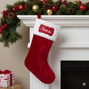 May include: A festive red Christmas stocking with a white cuff, bearing the name "Anna" on a red label. It hangs from a white mantelpiece adorned with a garland of greenery, red berries, and Christmas ornaments.
