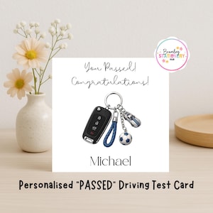 May include: A square greeting card featuring the text You Passed! Congratulations and the name Michael. The centre displays a black car key fob, a blue braided leather strap, a silver car charm, and a football charm on a ring.