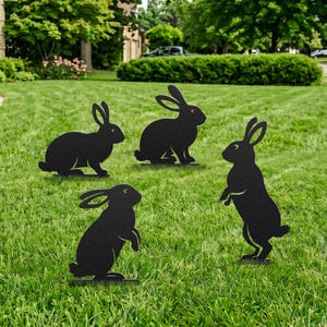 Peut inclure: Quatre décorations de jardin en métal noir en forme de lapin sont exposées sur une pelouse verte. Les lapins sont dans différentes poses, dont debout et assis. La pelouse est entourée d'arbres et de buissons.