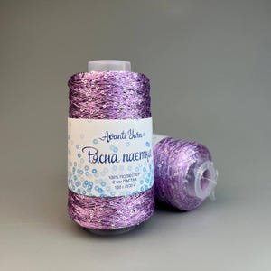 May include: A spool of purple yarn interwoven with small, reflective silver sequins. A white paper label wraps around the centre, featuring the text Avanti Yarn, 100% polyester, 2 mm, 100 g, and 530 m. A second, smaller spool of the same yarn lies behind it.