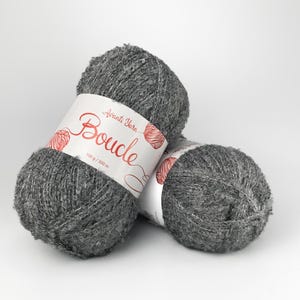 May include: Two skeins of dark gray textured yarn with a looped surface. Each skein features a white paper label with the red text Avanti Yarn Boucle and the measurement 100 g / 300 m printed on the front, indicating the weight and length.