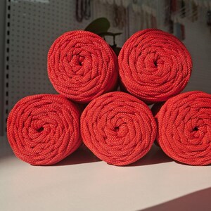 May include: Five spools of vibrant red macrame cord arranged in a pyramid formation. Each spool features a tightly wound, braided texture with a consistent circular cross-section, showcasing the uniform thickness and bright red color of the material.