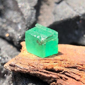 May include: A vivid, emerald-green gemstone, rectangular in form. The gem sits on a piece of weathered, brown wood, providing a natural contrast. The stone's facets reflect light, highlighting its clarity and colour. A close-up shot emphasises the gem's raw, unpolished beauty.