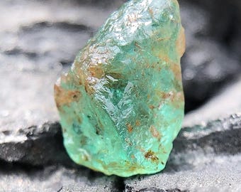 May include: A raw, uncut emerald gemstone with a translucent, glassy appearance. The gem has a vibrant green hue with hints of brown and is set against a dark, textured background. This natural emerald is a beautiful example of raw mineral beauty.