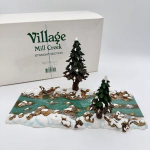 May include: A Department 56 Village Mill Creek straight section miniature display piece featuring a teal painted stream with brown rocks and white snow accents, accompanied by two dark green pine trees, presented with its original white box.