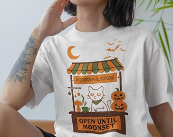May include: White t-shirt with a Halloween design. The graphic shows a cat, pumpkins, and the text "Potion & Chai" and "Open Until Moonset". The design uses orange, green, and brown colours.