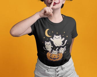 以下が含まれることがあります： ハロウィーンをテーマにしたグラフィックが特徴の黒いTシャツ。漫画の猫、幽霊、三日月、星が描かれています。猫は彫刻されたカボチャの中や周りにいます。背景は無地のオレンジ色です。