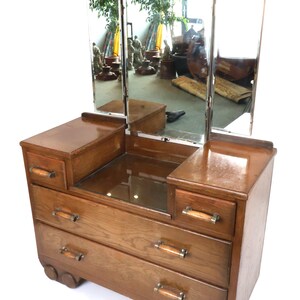 May include: A vintage wooden dressing table with a tri-fold mirror. The dressing table has three drawers and two side compartments with small drawers. The mirror is framed in a copper-coloured metal. The dressing table is made of light brown wood.