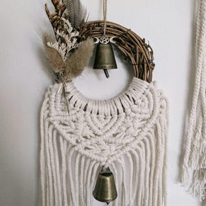 May include: A wall hanging featuring a circular grapevine wreath adorned with dried grasses, feathers, and a small silver-toned sword charm. It includes two brass-coloured bells, one with a pentacle symbol, and off-white macrame cord fringe.