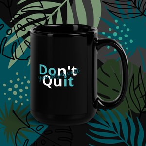 Op de afbeelding: Een zwarte koffiemok met de tekst "Don't Quit" in witte en blauwgroene letters. De mok heeft een grote handgreep en staat tegen een achtergrond van blauwgroene en groene tropische bladeren.