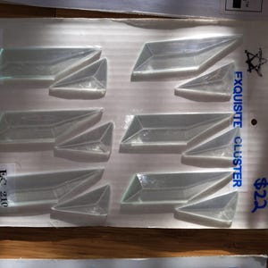 May include: A package of eight clear, geometric-shaped decorative pieces. The pieces are arranged on a white cardboard backing with the text "Exquisite Cluster" and "EC 210". The package also has the text "R.A.C. INC."