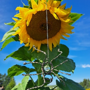 May include: A handcrafted wire spider pendant with a faceted crystal center hangs in front of a large sunflower. The spider is made of twisted metal wire and suspended from a delicate chain. The sunflower has bright yellow petals and a dark brown center, set against a blue sky.