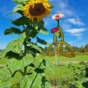 May include: A tall sunflower with a large yellow bloom and a brown center stands in a field. A stained glass flower holder with pink and red flowers hangs in front of the sunflower. The sky is blue.