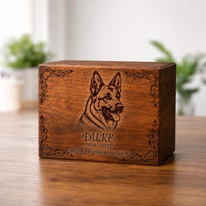 Op de afbeelding: Een rechthoekige houten doos met een donkerbruine afwerking, voorzien van een lasergegraveerd portret van een Duitse herder. De tekst luidt DUKE, 2010-2024, en You left paw prints on our hearts, omringd door decoratieve krullen en hartjes.