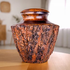 May include: A handcrafted urn with a textured, bark-like exterior. The urn is primarily brown with reddish-brown and dark brown accents. The lid is a darker shade of brown, creating a contrast. The urn is designed to hold cremated remains.