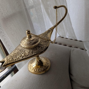 Puede incluir: Una lámpara de genio dorada con intrincados patrones grabados. La lámpara presenta un asa curva con una cadena, un pico y una tapa decorativa. La lámpara se exhibe sobre un cojín gris.