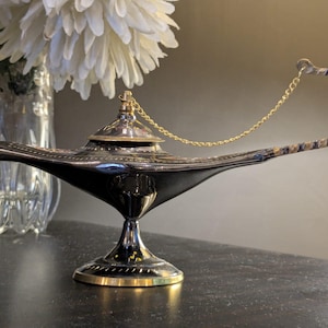 May include: A decorative genie lamp with a black body and a silver-coloured top and base. The lamp features a curved handle with a patterned design and a gold-coloured chain. The lamp is displayed on a dark surface.