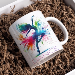 May include: White ceramic mug featuring a colorful watercolor design of a dancing figure with the word "DANCE" in purple. The mug is inside a white box filled with brown paper filler.