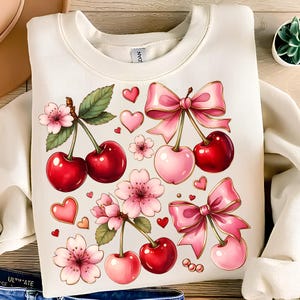 May include: Cream-coloured sweatshirt with a cherry design. The design features red and pink cherries, pink bows, hearts, and floral accents. The graphic is a whimsical and feminine illustration.