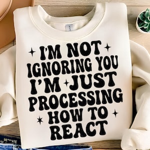 May include: Cream-colored sweatshirt with the text "I'M NOT IGNORING YOU I'M JUST PROCESSING HOW TO REACT" in black, stylized font. The text is surrounded by star shapes. The sweatshirt is folded.