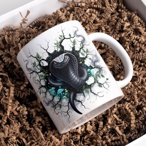 May include: White ceramic mug with a black cobra design breaking through a cracked surface, with green and blue details. The mug has a white handle and is presented in a box with brown packing material.